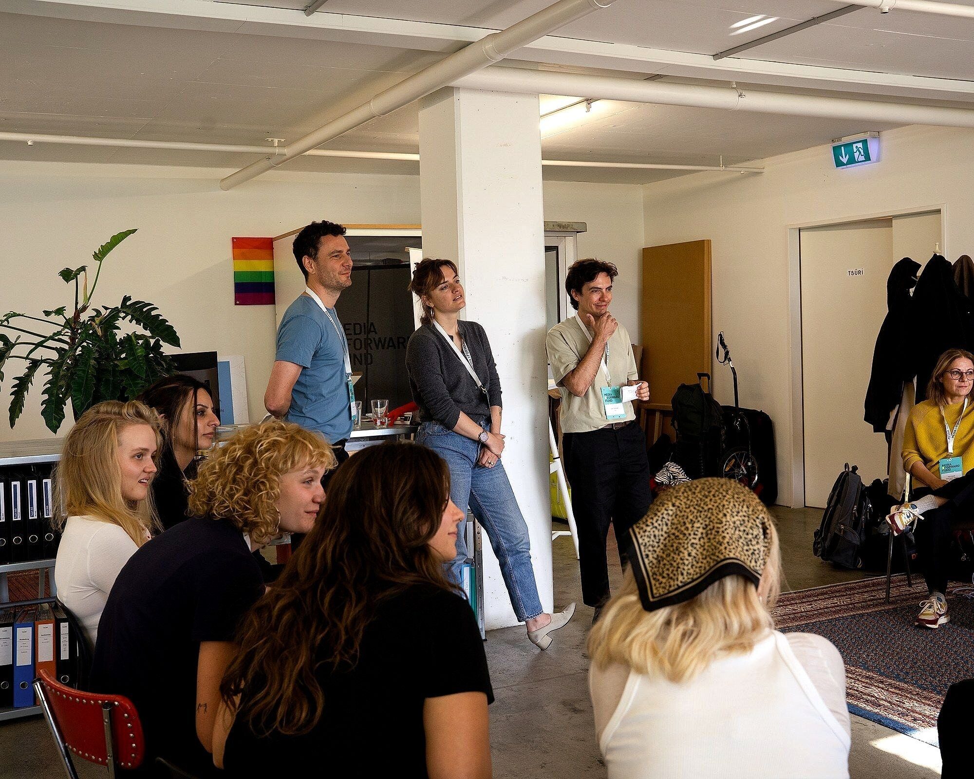 Gruppe von Personen mit Namensschildern sitzt und steht in einem Büroraum, im Hintergrund eine Regenbogenfahne an der Wand.